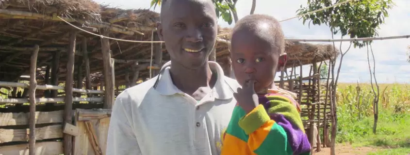 A man holding a child in front of a hut.