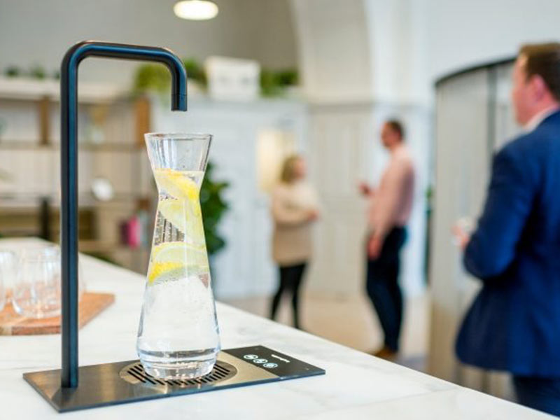 Dispense A glass of water on a counter with people standing around.
