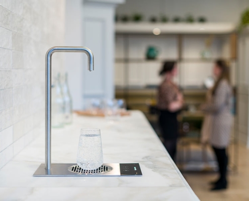Gallery-1 A woman is standing next to a water dispenser in a kitchen.